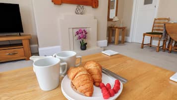 A dining table with croissants and raspberries, two white mugs, a fireplace with flowers, a television, and a mirror.