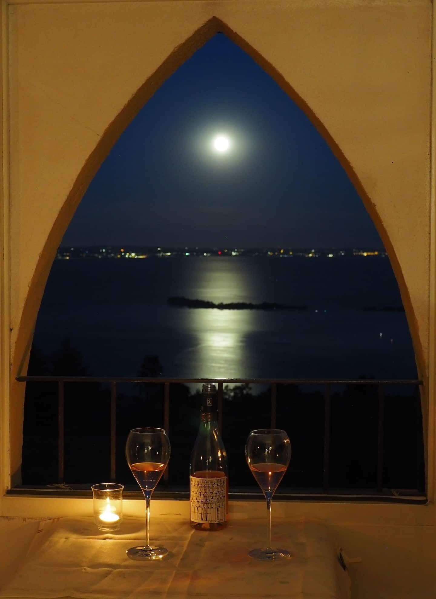 Een raam met uitzicht op een meer in maanlicht en lichtjes van de stad, een fles wijn en twee wijnglazen op een tafel.