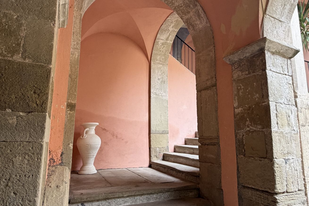 Un corridoio in pietra con archi, un vaso e una scala.