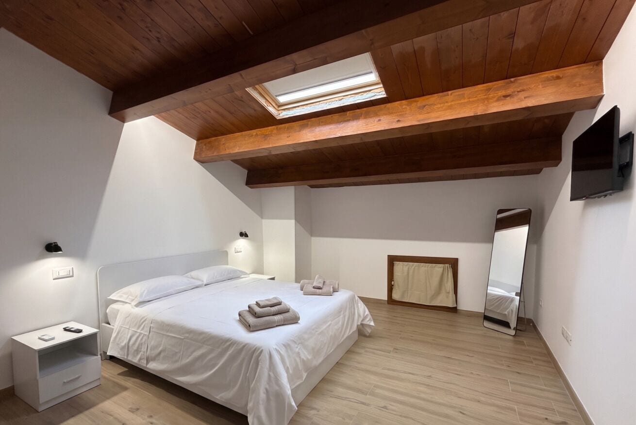 Una camera da letto con soffitto spiovente, una finestra a lucernario, un letto con biancheria bianca, un comodino con lampada, uno specchio e un pavimento in legno.