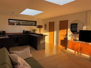 A modern kitchen with a central island, a dining area with a sofa, and a TV on a wooden cabinet. - Birdsong (Penzance)