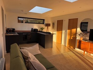A modern kitchen with a central island, a dining area with a sofa, and a TV in a wooden cabinet. - Birdsong (Penzance)