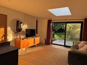 A modern living room with a flat-screen TV, a wooden cabinet, a round mirror, and a sliding glass door leading to a patio with outdoor furniture. - Birdsong (Penzance)
