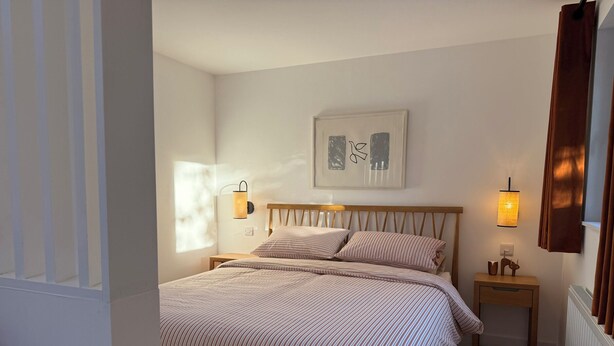 Une chambre à coucher avec un lit, deux tables de chevet, une lampe fixée au mur, un tableau encadré et une fenêtre avec des rideaux.
