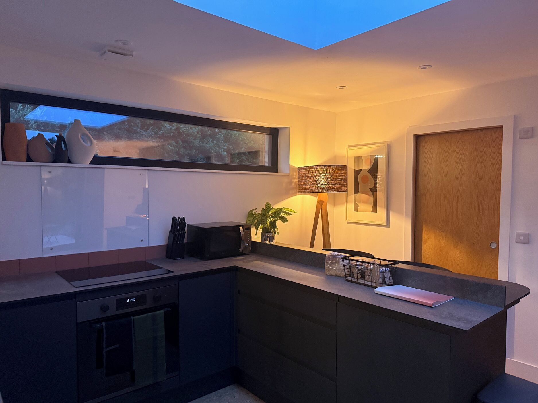 A modern kitchen with a built-in microwave, a sink, and a small oven. There is a window above the sink, a potted plant, and a framed artwork on the wall.