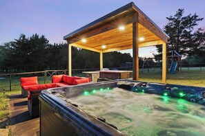 Un bain à remous avec une terrasse en bois et des coussins rouges, un espace salon couvert et, au fond, une aire de jeux.