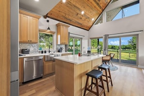 A modern kitchen with wooden cabinets, a stainless steel dishwasher, and a large island with bar stools. - Luxury Ranch Retreat, Hot Tub, Private Fishing Pond, Longhorns, Sleeps 6 (Bonham)