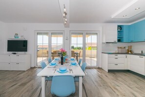 Een moderne keuken met witte kasten, een eethoek met een witte tafel, blauwe stoelen en een televisie op een wit meubel.