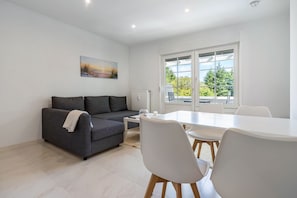 Ein modernes Wohnzimmer mit grauem Sofa, weißem Esstisch und Stühlen, einem Bild an der Wand und einem großen Fenster mit Blick.