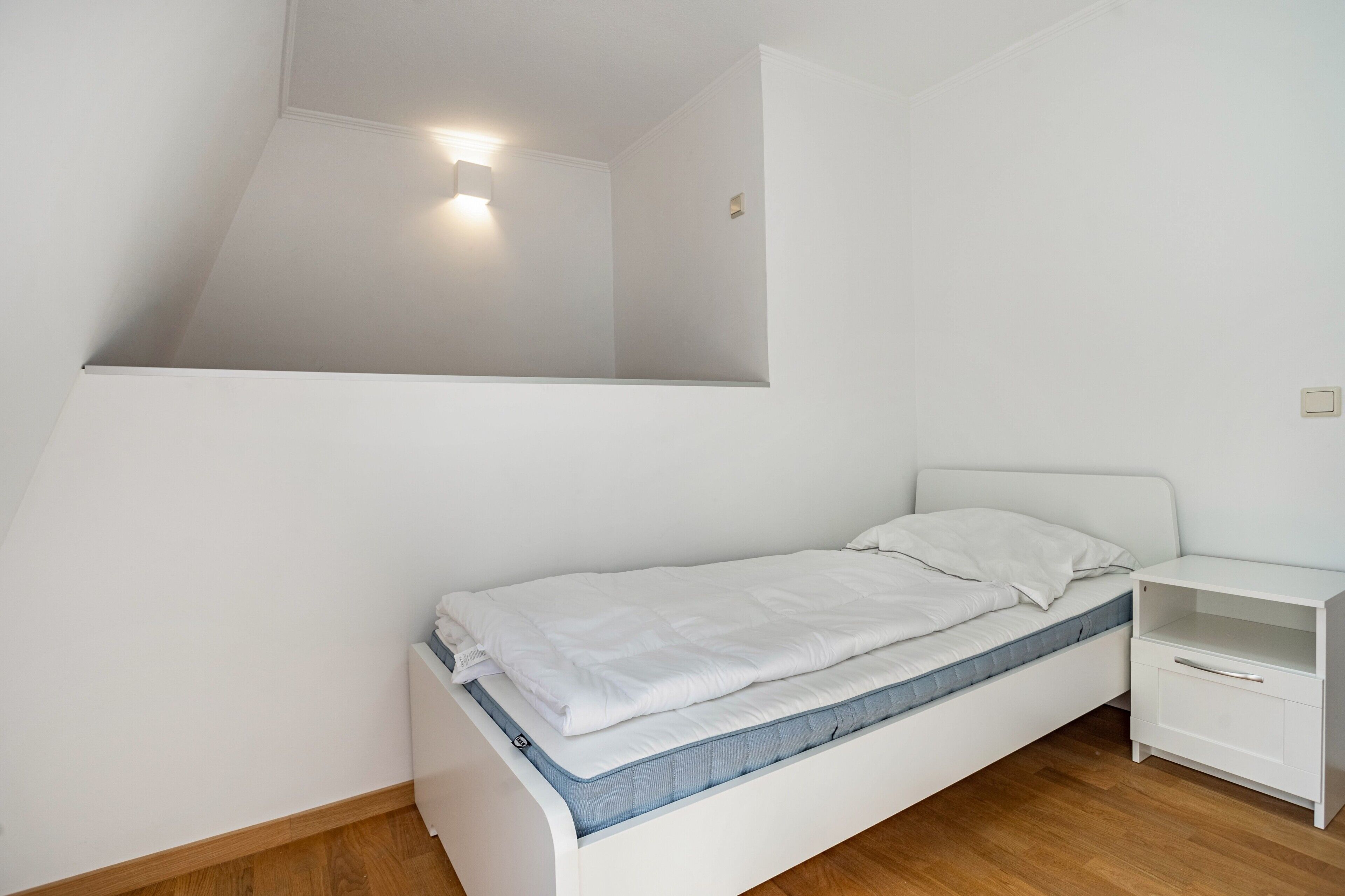 A small, white, single-bed room with a sloped ceiling, a bedside table, and a wall-mounted light fixture.