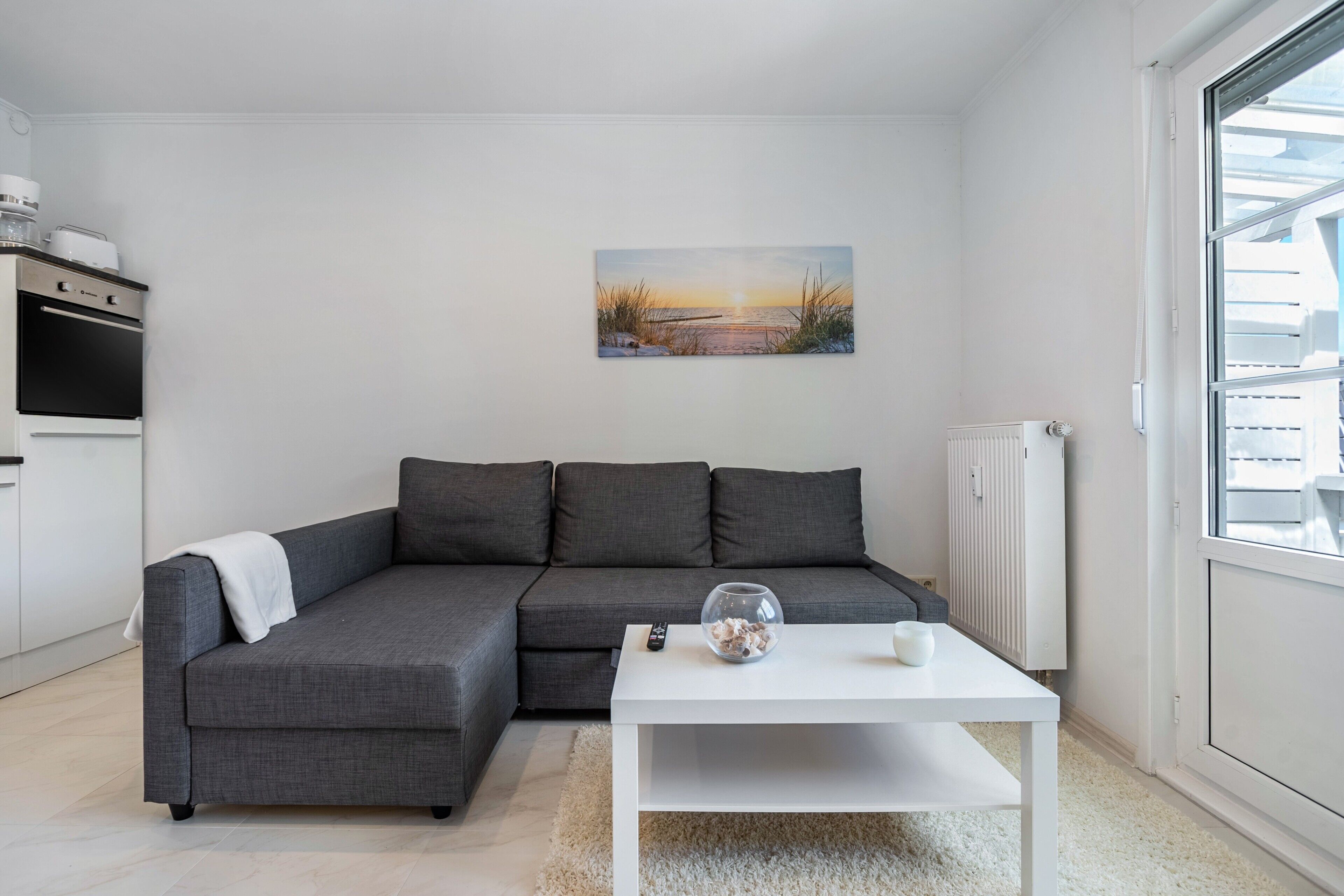 Un salon moderne avec un canapé d’angle gris, une table basse blanche et un tableau représentant un coucher de soleil sur l’eau.
