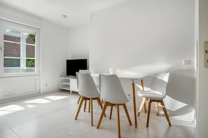 Un comedor moderno con una mesa y sillas blancas, un televisor sobre un mueble blanco y una ventana con vista a una casa.