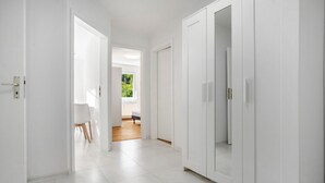 Um corredor com paredes e portas brancas, que dá para um quarto com cama e janela.