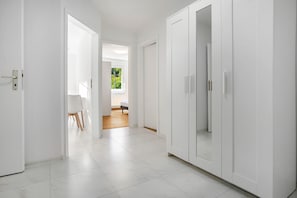 Um corredor com paredes e portas brancas, que dá para um quarto com cama e janela.