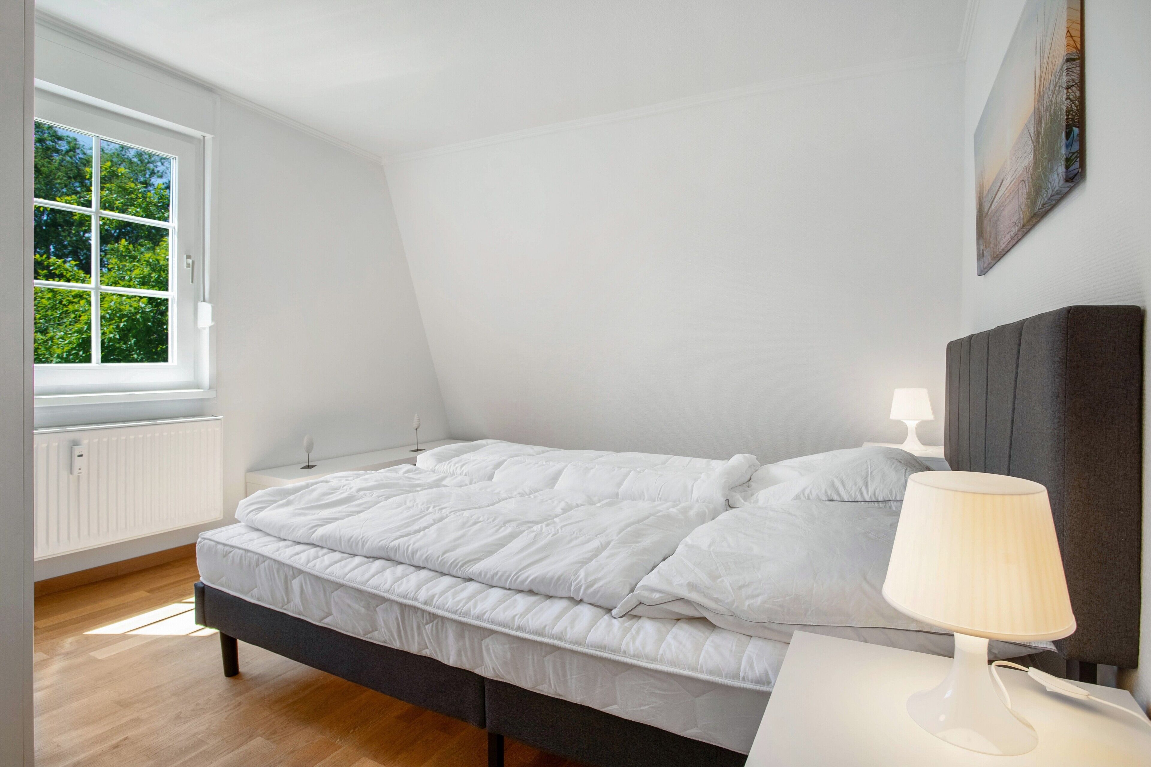Un dormitorio con cama, lámpara de noche, ventana y un cuadro en la pared.