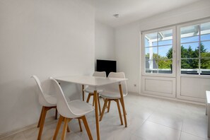 Una habitación con una mesa y sillas blancas, un televisor de pantalla plana y un ventanal con vistas a árboles.