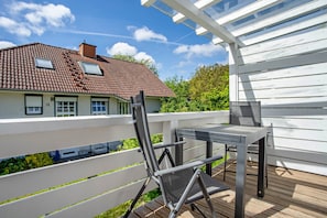 Um deck de madeira com mesa e cadeiras, um corrimão branco e uma casa com telhado de cerâmica ao fundo.