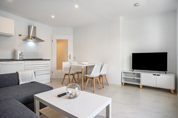 Uma sala de estar moderna com sofá, mesa de centro, televisão, área de jantar com cadeiras e cozinha com fogão e armários.