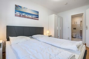 Un dormitorio con cama, mesita de noche, lámpara de pared, una puerta que da a un baño y un cuadro con una playa.