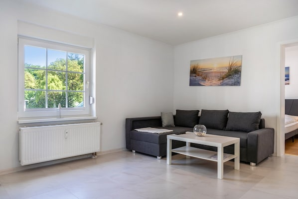 Ein modernes Wohnzimmer mit einem grauen Sofa, einem weißen Couchtisch, einem Bild an der Wand und einem Fenster mit Blick auf Bäume.
