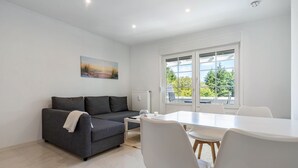 A modern living room with a grey sofa, white dining table, and chairs, a painting on the wall, and a large window with a view. - Apartment 'Whg 2.4 1.OG Rechts Vorn' with Balcony (Putbus)