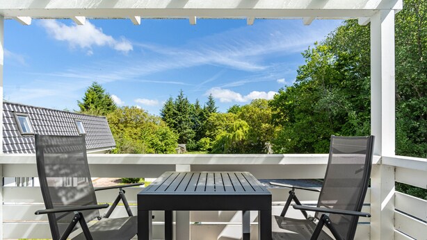 A covered outdoor area with a table and two chairs, surrounded by a white railing, overlooking a residential area with trees and a house. - Apartment 'Whg 2.4 1.OG Rechts Vorn' with Balcony (Putbus)