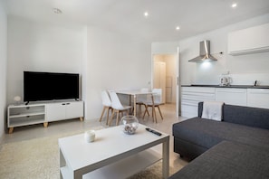 Un salon moderne doté d’un téléviseur à écran plat, d’une table à manger avec des chaises, d’un canapé gris et d’un coin cuisine avec des placards et une hotte aspirante.