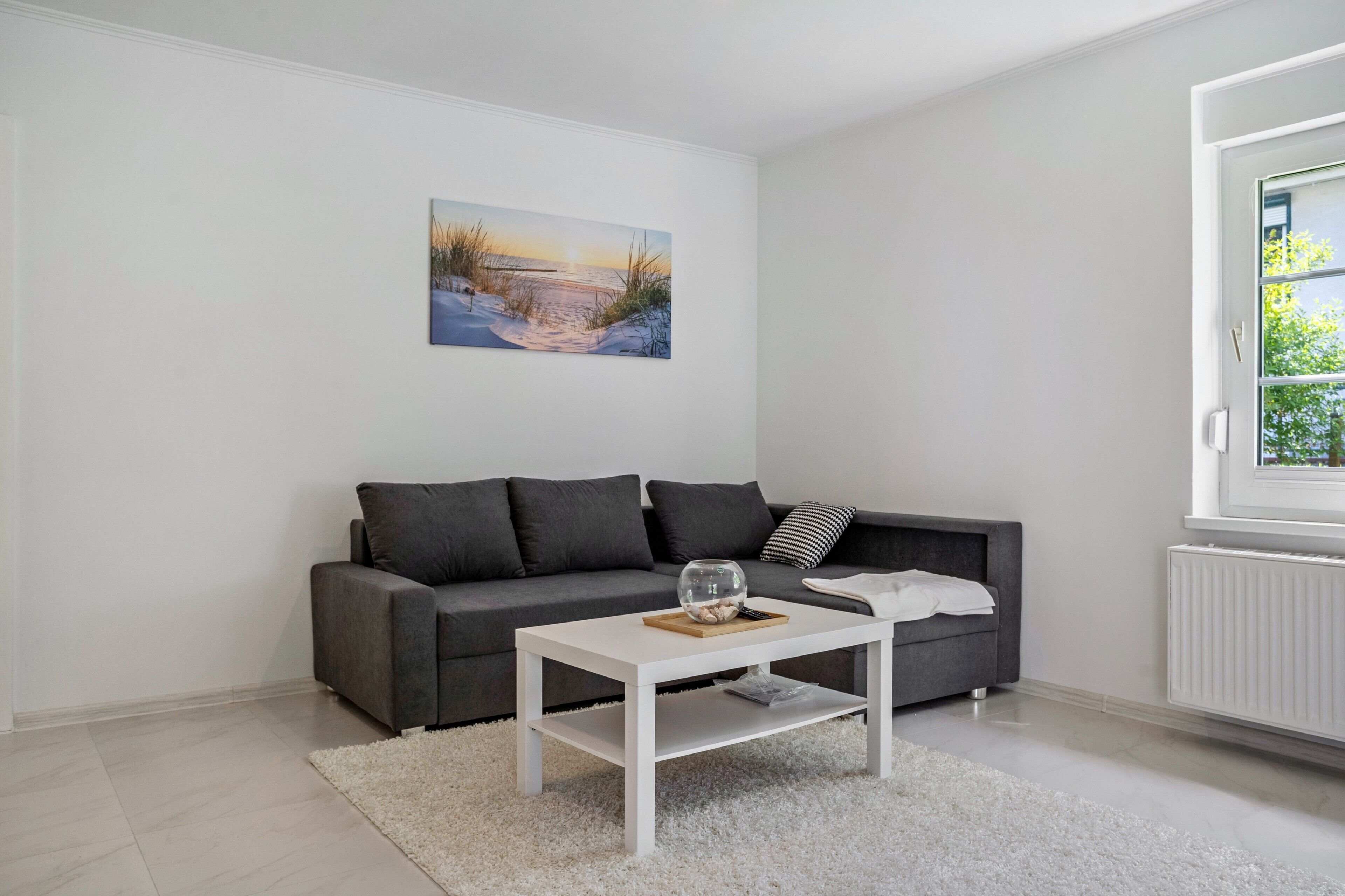 Ett modernt vardagsrum med en grå soffa, ett vitt soffbord och en tavla som föreställer en strandscen.