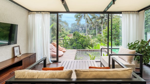 A balcony with a wooden floor, a sofa, a TV, and a view of a garden. - Villa Oasis (Ubud)