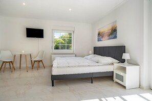 Un dormitorio con cama, televisor, ventana, un cuadro y un escritorio con sillas.