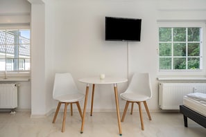Una habitación con pared blanca, un televisor de pantalla plana montado sobre una pequeña mesa redonda con dos sillas blancas, una ventana que da a vistas de vegetación y un radiador en el suelo.