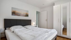 A bedroom with a bed, a nightstand, a bedside table, a wardrobe, and a painting on the wall. - Apartment 'Whg 2.8 1.OG Rechts Hinten' with Balcony (Putbus)
