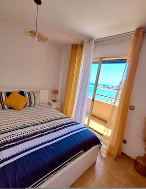 A bedroom with a bed, a nightstand, a lamp, and a view of the sea through a balcony. - Beachfront 3BR Condo w/ Pool Views | Sleeps 10 (La Manga)