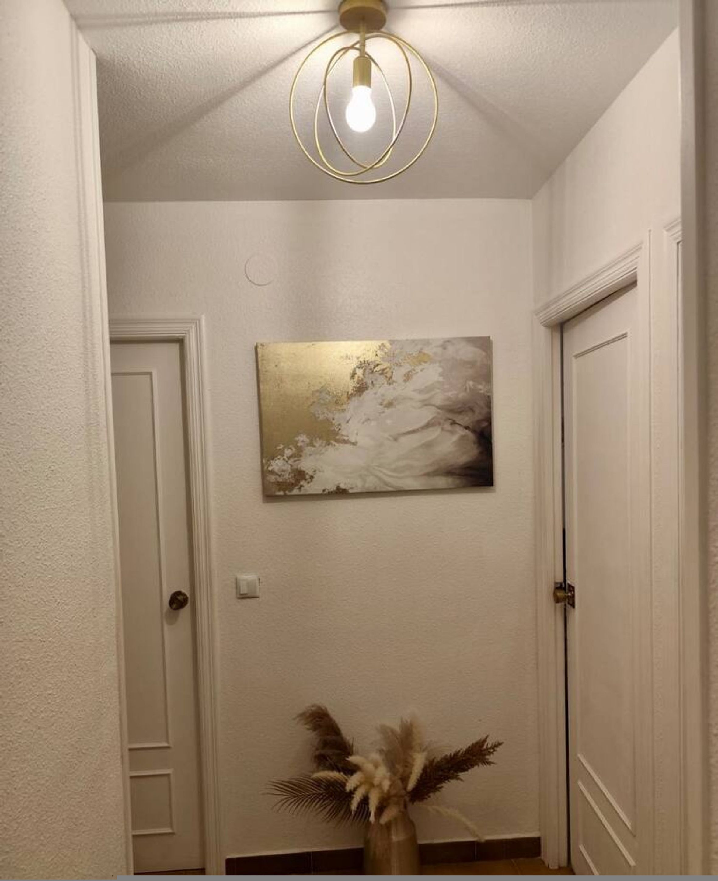 A hallway with a ceiling light fixture, a wall-mounted abstract painting, and a vase with dried plants.