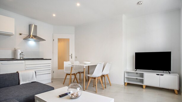 Een moderne woonkamer met een bank, een salontafel, een televisie, een eethoek met stoelen en een keuken met een fornuis en kastjes.