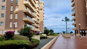 Un hotel de varios pisos con balcones, un camino pavimentado y zonas ajardinadas con arbustos y flores.