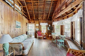 Interior de una cabaña de madera con sofá, sillas, una mesa y un televisor.