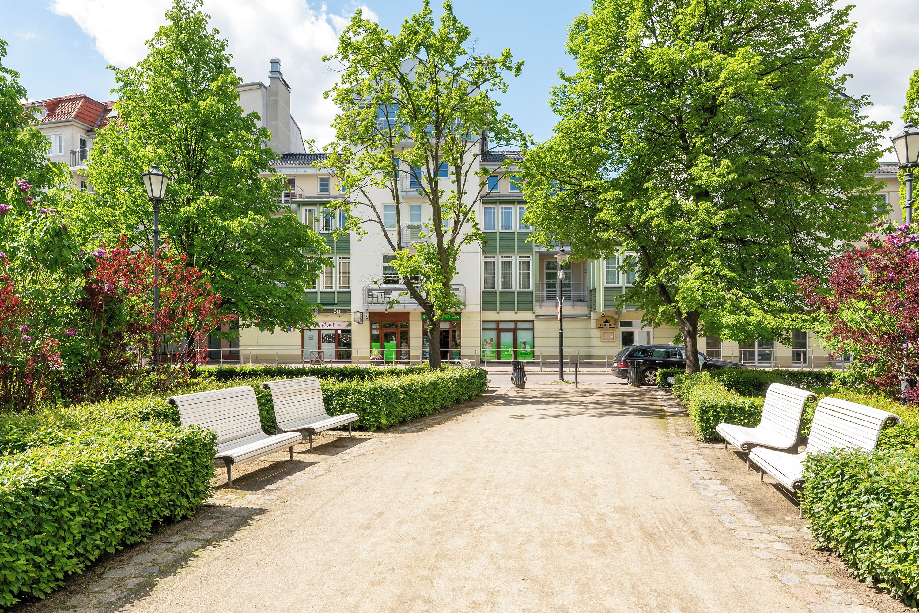A paved walkway with benches, surrounded by greenery and buildings with visible signage.
