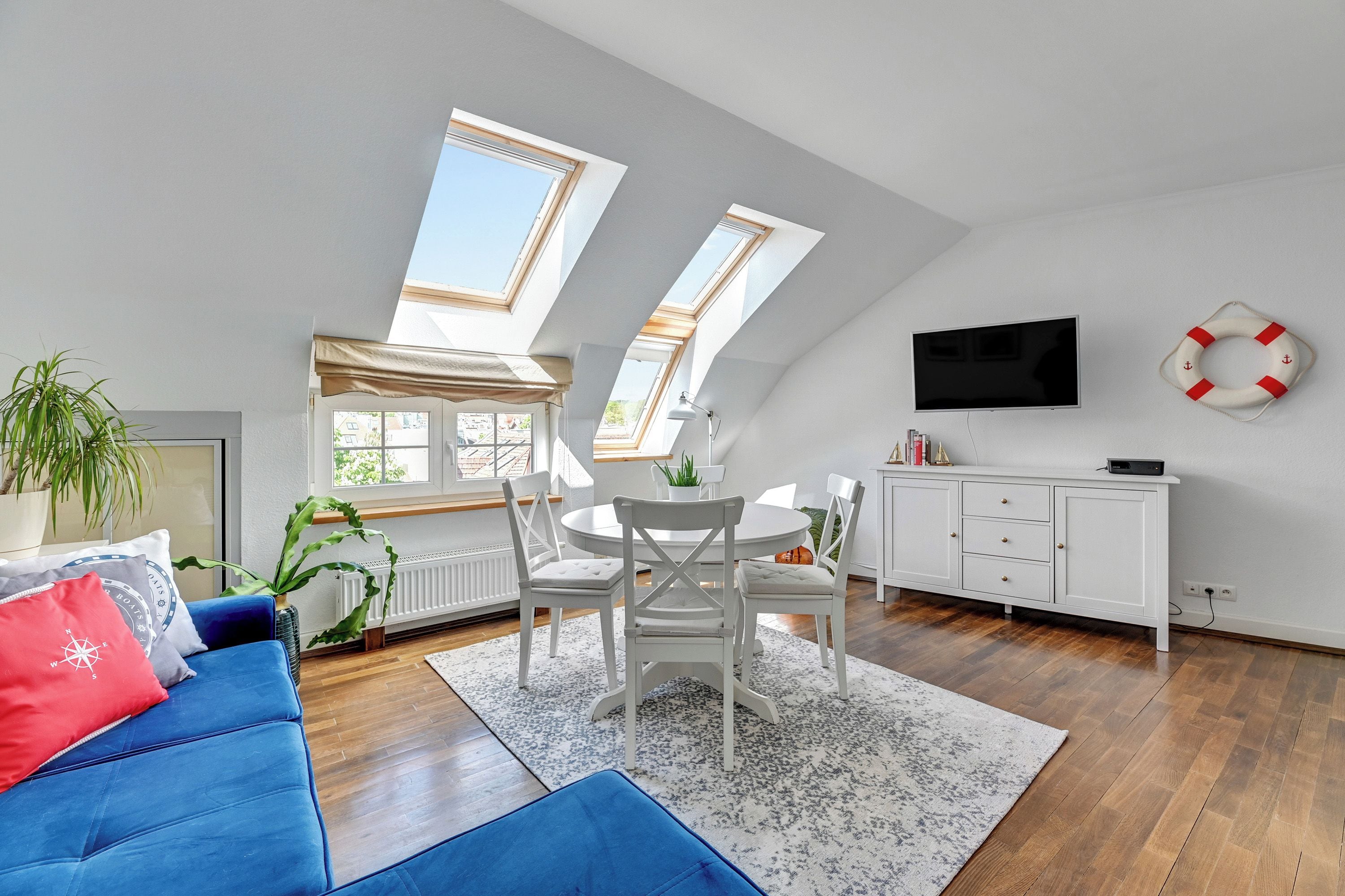A modern living room with a blue sofa, a dining table with chairs, a white cabinet, and a television on the wall.