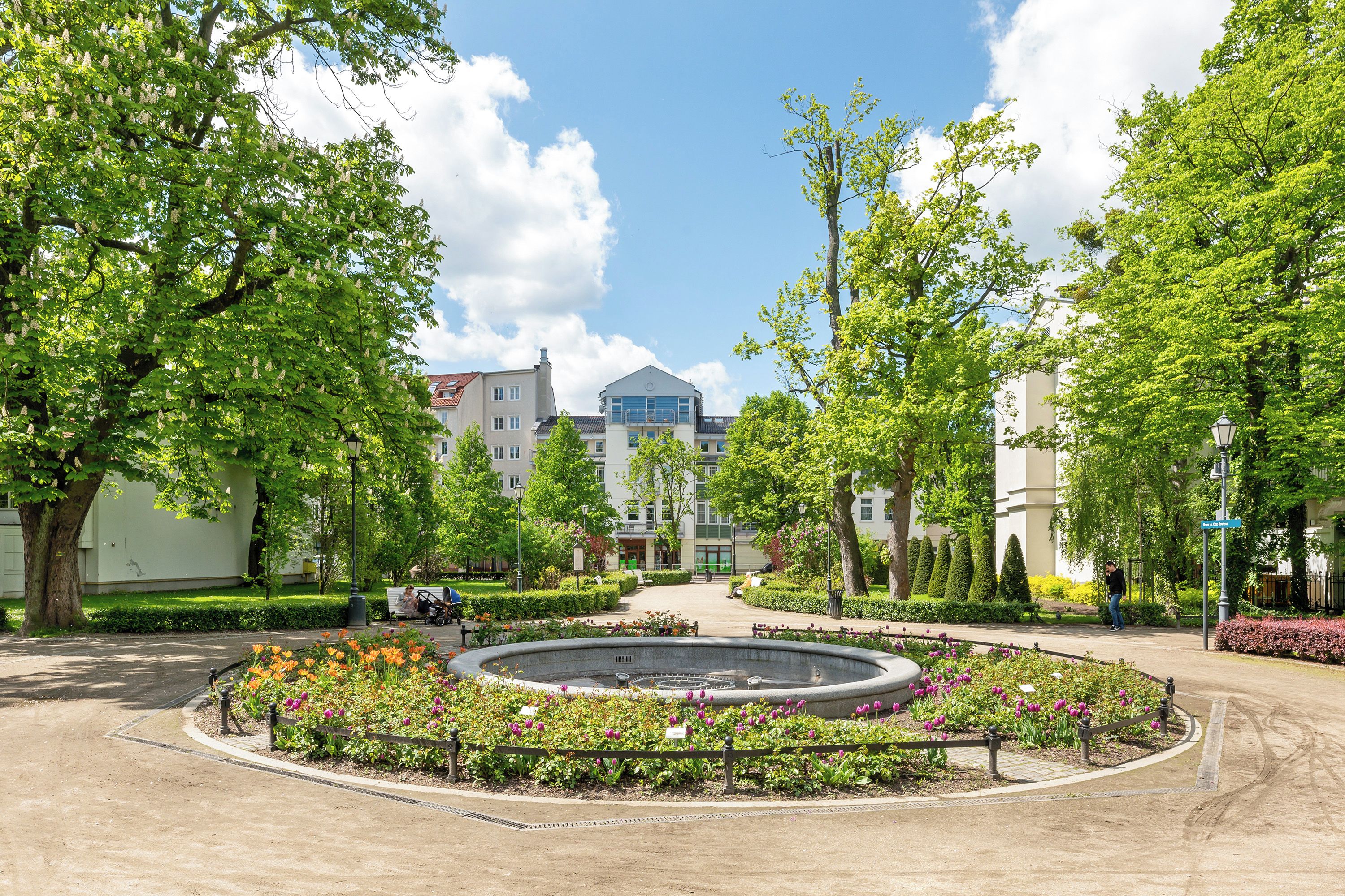 Une cour aménagée avec une fontaine centrale, entourée de parterres de fleurs et d’arbres, qui mène à un bâtiment doté de nombreuses fenêtres et d’un toit bleu.