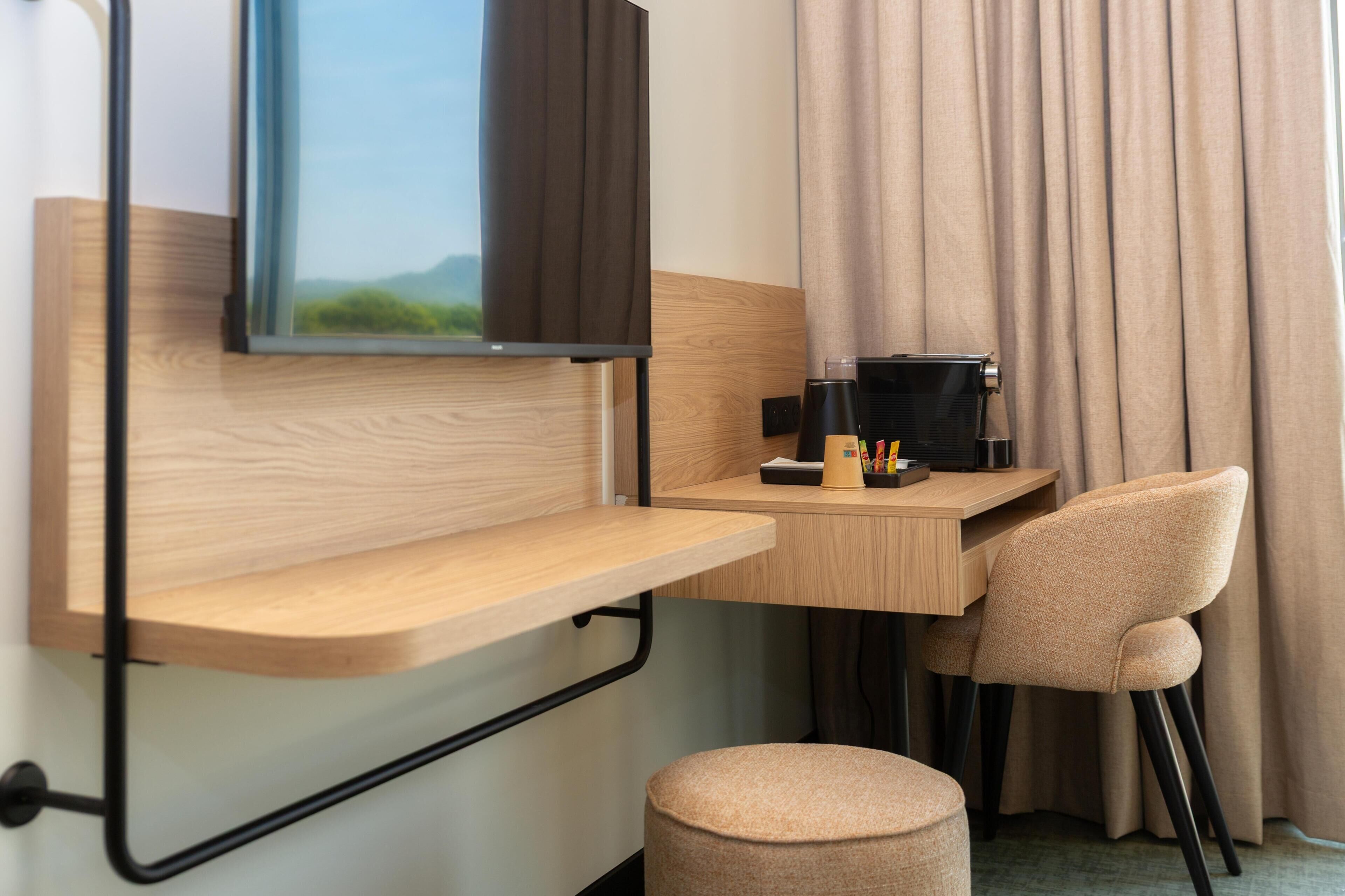 Une chambre d’hôtel de taille modeste, dotée d’un bureau en bois, d’une chaise, d’un petit pouf, d’un téléviseur fixé au mur et d’une fenêtre avec des rideaux.