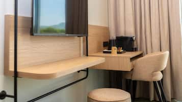 Une chambre d’hôtel de taille modeste, dotée d’un bureau en bois, d’une chaise, d’un petit pouf, d’un téléviseur fixé au mur et d’une fenêtre avec des rideaux.