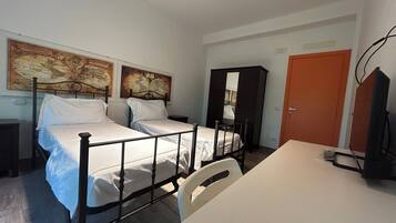 A bedroom with two beds, a desk, a chair, a wardrobe, and two wall-mounted artworks.