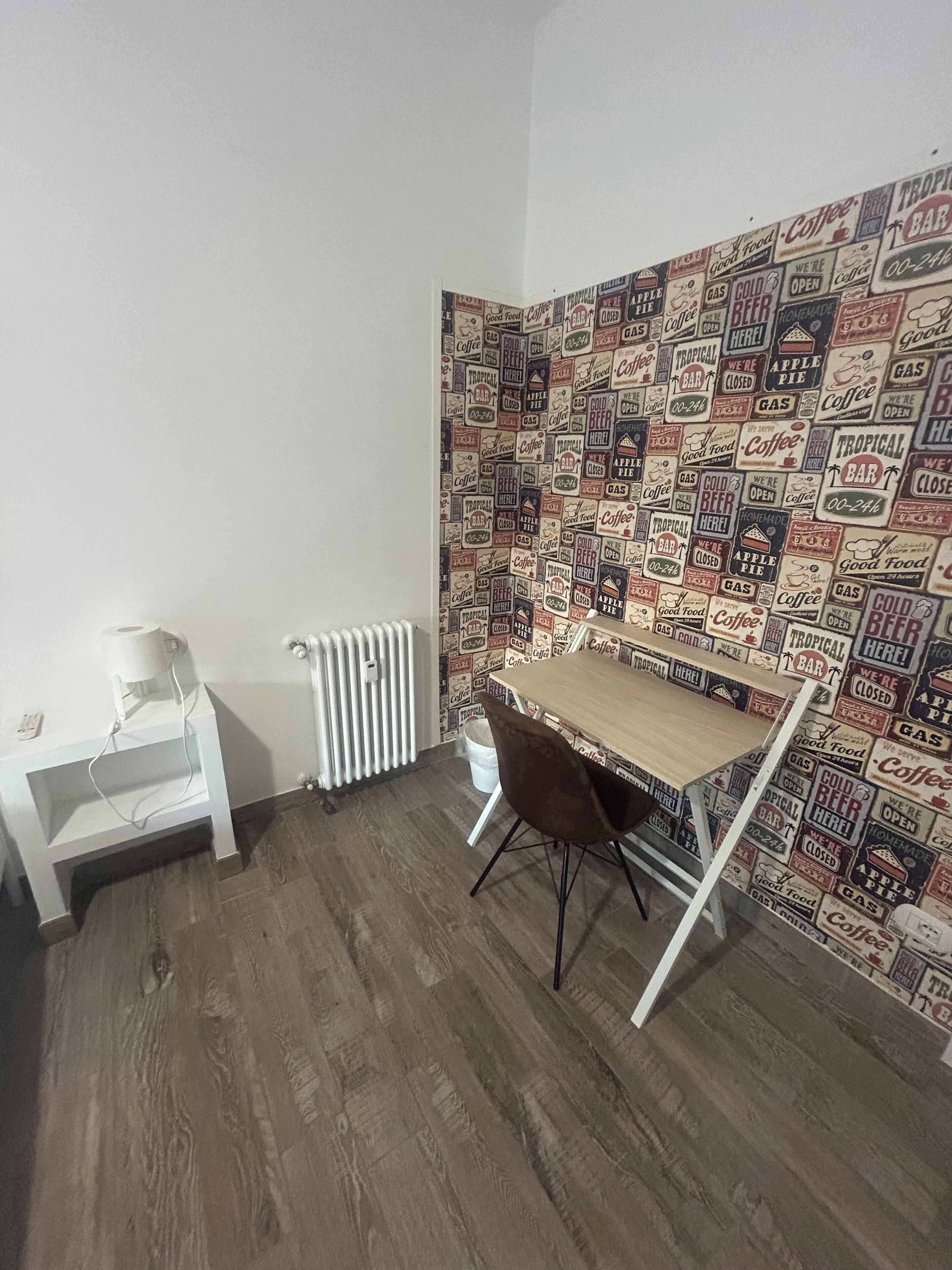 Una habitación con suelo de madera, un radiador blanco, un escritorio pequeño con una silla y una pared decorada con carteles de estilo vintage.