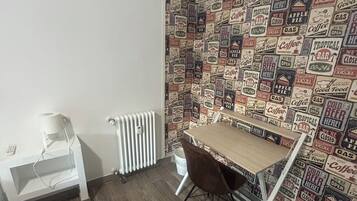 Una habitación con suelo de madera, un radiador blanco, un escritorio pequeño con una silla y una pared decorada con carteles de estilo vintage.