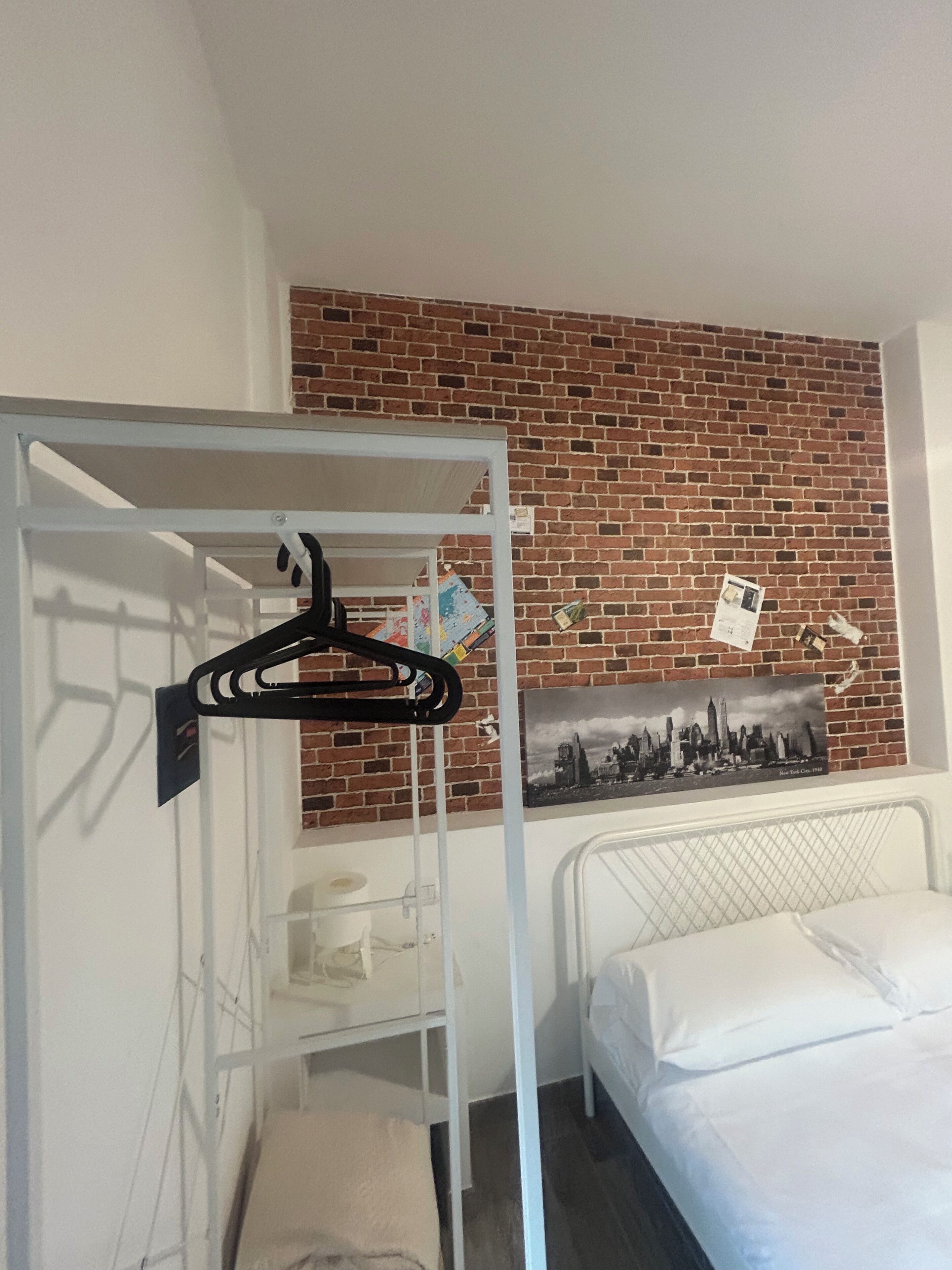 A room with a bunk bed, a white metal wardrobe, a brick wall, and a cityscape picture.