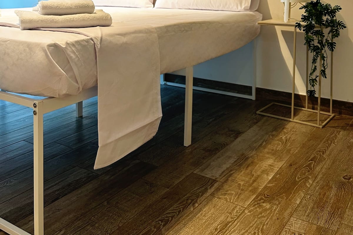 Una cama bien hecha con sábanas blancas, cabecera de madera, mesita de noche con lámpara y una planta en maceta.