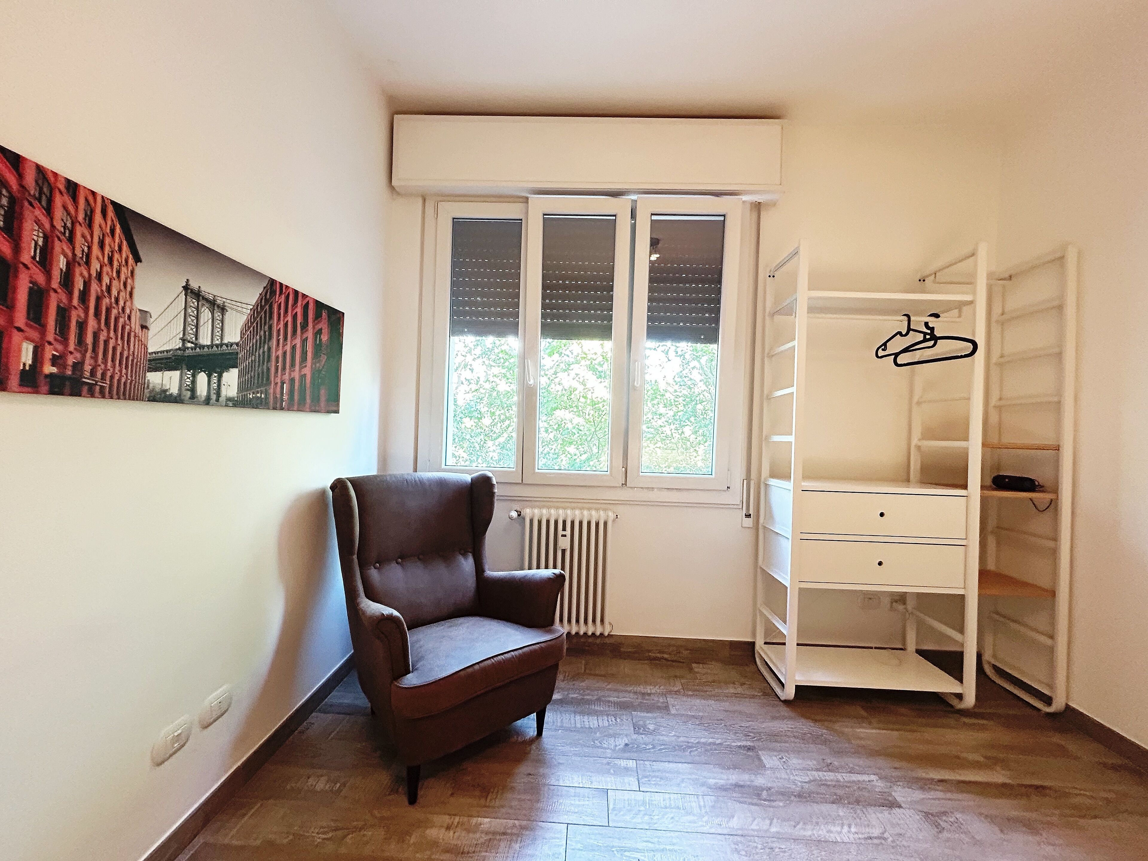 Une pièce avec un fauteuil marron, une étagère blanche, un radiateur et une fenêtre donnant sur un pont.