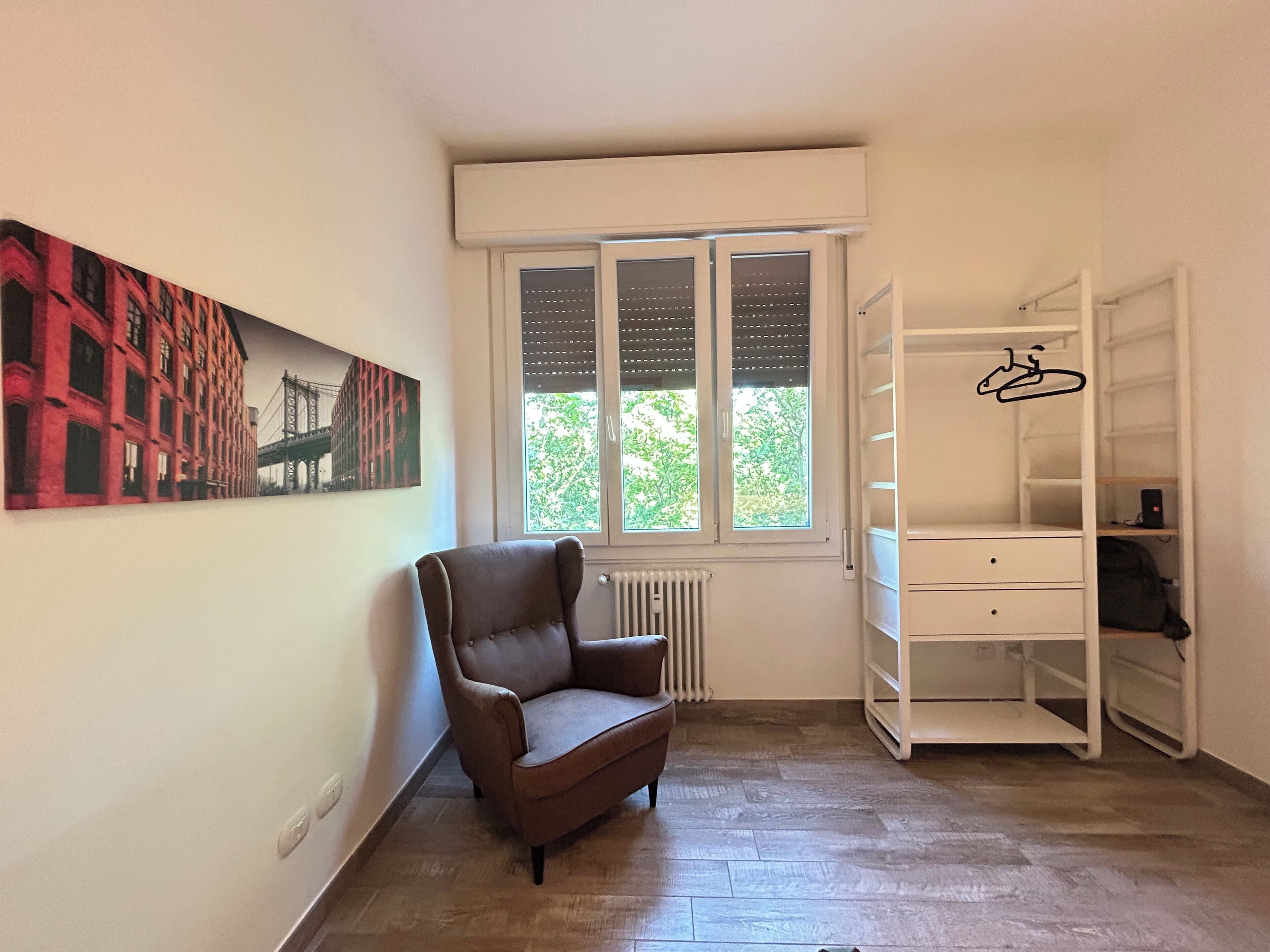 A room with a radiator, a window with blinds, a chair, a white shelving unit, and a painting of a bridge.