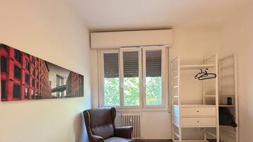 A room with a radiator, a window with blinds, a chair, a white shelving unit, and a painting of a bridge.